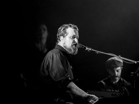 Black and white head shot of bearded man playing piano and singing into microphone