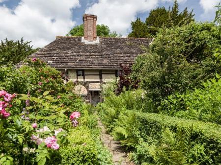 The Priest House stands in a traditional cottage garden. Greenery with pink flowers in front of the house.