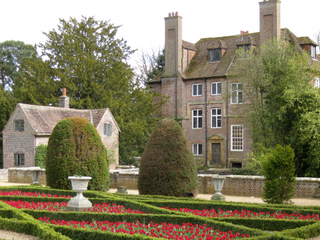 Photo of the house at Groombridge Place.
There is red flowers in patches in front on the house which is interwind with bushes.