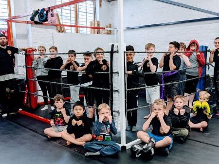 A group of children standing in a boxing ring and sitting around it with two instructors