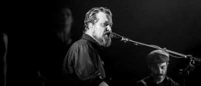 Black and white head shot of bearded man playing piano and singing into microphone