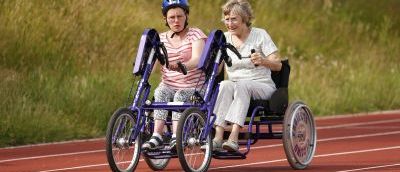 Two people riding on conjoined bike, an elderly person with a young person who is wearing a helmet.