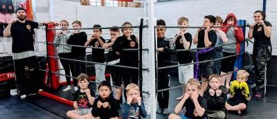 A group of children standing in a boxing ring and sitting around it with two instructors